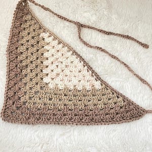 May include: A crocheted triangular scarf in shades of brown and white. The scarf has a fringe on the bottom edge and is tied with a long cord.