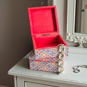 May include: Open wooden jewelry box with a red interior and a colorful floral pattern on the exterior. A silver chain with large links rests on the box. The box is stacked on top of another box with the same design.
