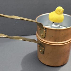May include: A double boiler set featuring two stacked copper pots with brass handles. The top pot contains a silver-toned metal insert topped with a small, yellow, molded plastic chick figure that serves as a handle or decorative knob.