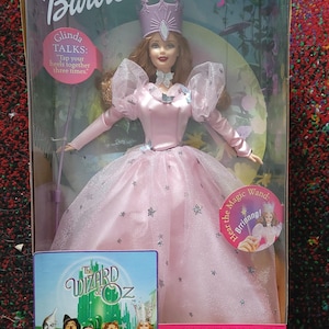May include: Barbie doll of Glinda from The Wizard of Oz, in a pink gown with a crown. The doll is in a clear plastic box with the Barbie logo and the words "Glinda Talks". A small box shows the movie characters.