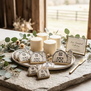May include: A display of barn-shaped cookies with black and white details, arranged on a white plate. Four small, round cakes are in the background. A gold-framed sign reads "First Farmstead Collection". A gold knife rests on the stone surface.