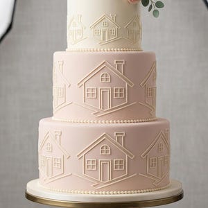 以下が含まれることがあります： 家をモチーフにした3段のケーキで、花が飾られています。下の2段は薄いピンク色で、上の段は白色です。ケーキは金の台の上に置かれています。ケーキには家のデザインが施されています。