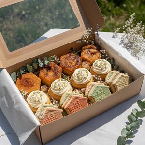 以下が含まれることがあります： 白いフロスティングと緑色のアクセントが付いたカップケーキ、六角形のケーキ、家型のクッキーなど、焼き菓子が詰められたギフトボックス。ボックスは白い紙で裏打ちされ、緑と花の小枝で飾られています。ボックスには透明な窓が付いています。