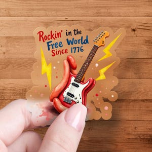 Peut inclure: Un autocollant représentant une guitare électrique rouge et blanche, tenue par un design en forme de main. Le texte "Rockin' in the Free World Since 1776" est affiché avec des éclairs jaunes. Le fond est une surface en bois.