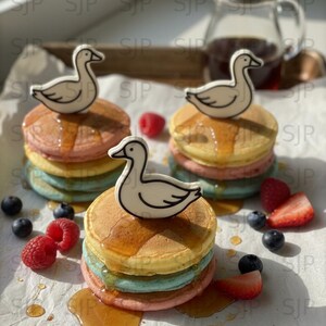 May include: Colorful pancakes with syrup, topped with white duck-shaped decorations. The pancakes are stacked with layers of pink, blue, and yellow. Fresh berries, including blueberries and raspberries, are scattered around the pancakes.