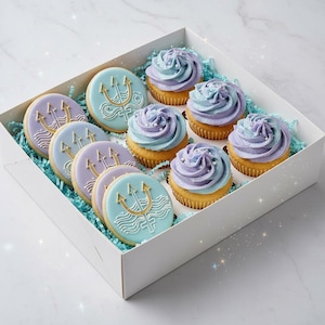 Può includere: Una scatola bianca piena di cupcakes e biscotti. I cupcakes hanno una glassa blu e viola. I biscotti rotondi presentano un design a tridente dorato. La scatola è foderata con sfilacci di carta blu.