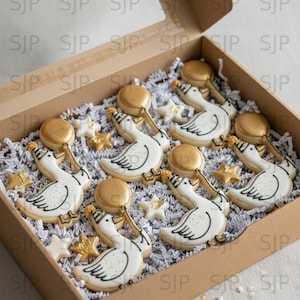 May include: A box of decorated cookies, featuring white swan shapes with black outlines and gold accents. The box also contains gold star-shaped cookies and gold-topped treats, all nestled in white paper shreds.