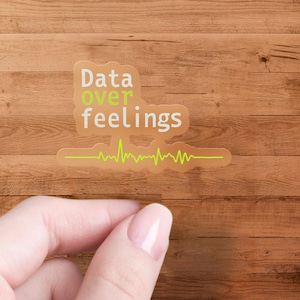 May include: A transparent sticker with the text "Data over feelings" in white and green, with a green heartbeat line below. The sticker is on a wooden surface.