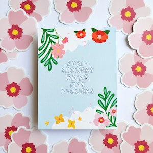 May include: A light blue card with the text "April Showers Bring May Flowers" in a white outline. The card is decorated with green leaves, pink and red flowers, and white clouds.