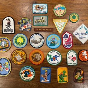 May include: A collection of vintage patches with various designs, including a tiger, a whale, and a cartoon turtle. The patches feature text such as "Cookies 1990", "Girl Scout Day Camp", and "Camp Winacka".
