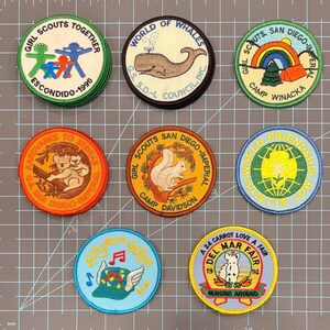 May include: A collection of nine colorful Girl Scout patches. The patches feature various designs, including a whale, a squirrel, a rainbow, and text such as "World of Whales" and "Del Mar Fair". The patches are round and have embroidered details.