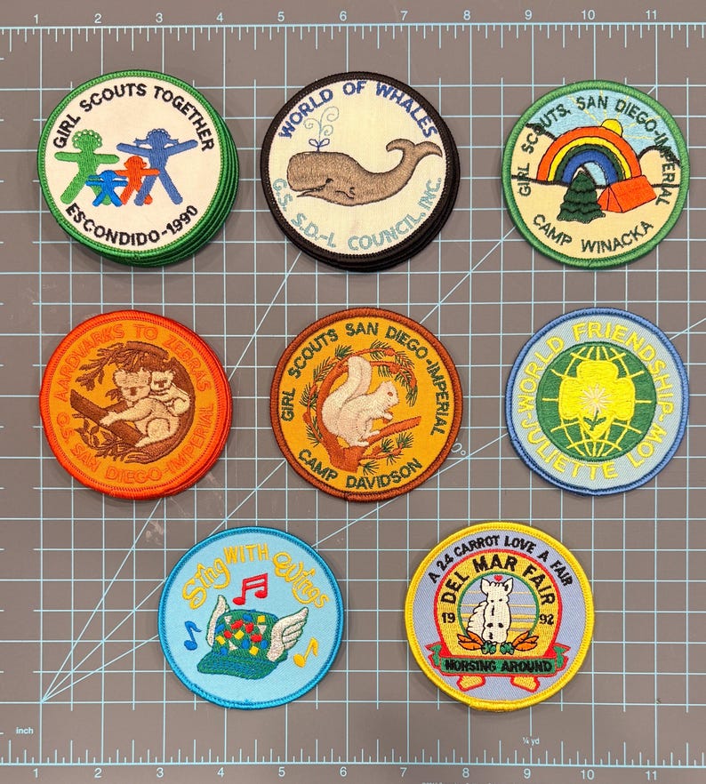 May include: A collection of nine colorful Girl Scout patches. The patches feature various designs, including a whale, a squirrel, a rainbow, and text such as "World of Whales" and "Del Mar Fair". The patches are round and have embroidered details.