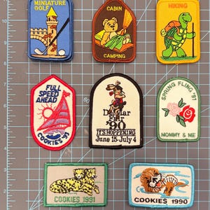 May include: A collection of embroidered patches with various designs. Themes include miniature golf, camping, hiking, sailing, and a fair. Some patches feature text like "COOKIES 1991" and "Del Mar Fair '90".