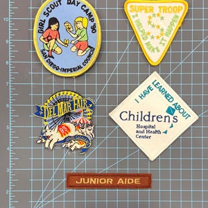 May include: A collection of embroidered patches. Includes a blue and yellow Girl Scout Day Camp patch, a yellow Super Troop patch, a Del Mar Fair patch, a Children's Hospital patch, and a Junior Aide patch.