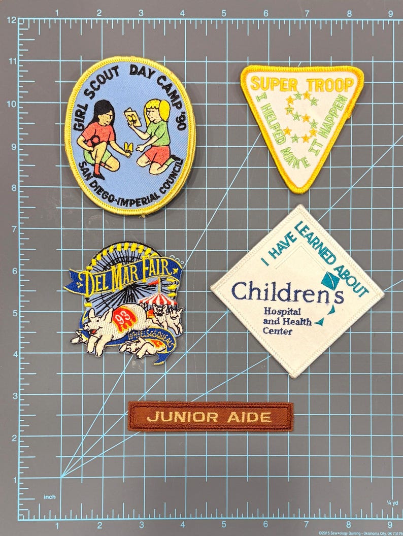 May include: A collection of embroidered patches. Includes a blue and yellow Girl Scout Day Camp patch, a yellow Super Troop patch, a Del Mar Fair patch, a Children's Hospital patch, and a Junior Aide patch.