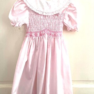 May include: A light pink dress with a white collar and short, puffed sleeves. The bodice features smocking detail with pink embroidery. The skirt is a full A-line shape, creating a classic, vintage look. The dress is hanging on a white hanger.