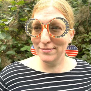 May include: A person wearing oversized round sunglasses and matching earrings, both featuring a patriotic red, white, and blue stars and stripes design. The person is wearing a black and white striped top.
