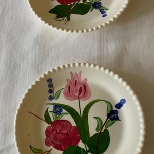 May include: Two off-white ceramic plates with scalloped edges. Each plate features a hand-painted floral design with red roses, pink tulips, green leaves, and blue accents. The plates are arranged on a white surface.