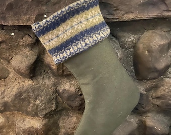 May include: A Christmas stocking with a green body and a patterned cuff. The cuff features a design of blue, tan, and white knit stripes. A dark blue loop is attached for hanging. The stocking is displayed on a stone fireplace.