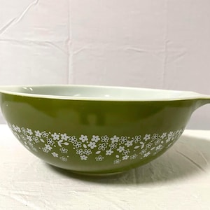 May include: A round, olive green mixing bowl featuring a white floral pattern around the exterior. The bowl has a smooth, glossy finish, a wide opening, and a small, pointed handle extending from one side of the rim, with a white interior.