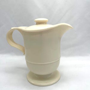 Puede incluir: Jarra de color crema con tapa y asa. La jarra tiene una base de pedestal y un borde decorativo. El asa es curva y la tapa tiene un pequeño pomo redondo. La jarra está hecha de un material opaco.