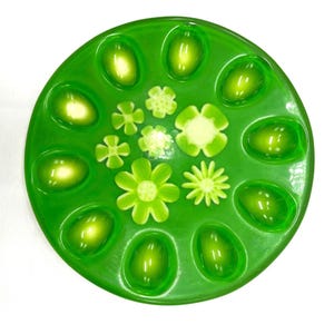 May include: A round, green decorative plate with a floral design. The plate features a glossy finish and is adorned with various flower shapes and oval accents in shades of green and white. The design is arranged in a circular pattern.