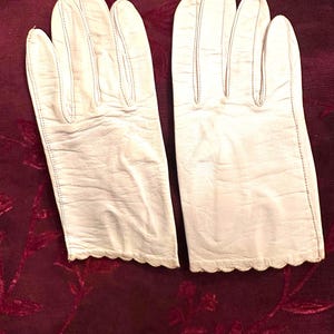 May include: A pair of white leather gloves featuring a smooth finish, visible stitching along the fingers, and a scalloped edge at the wrist. The gloves are constructed with a classic design, showcasing the natural texture of the material.