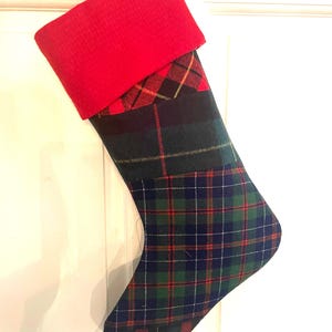 May include: A Christmas stocking with a red cuff and a plaid pattern in shades of green, blue, and red. The stocking is hanging against a white background, ready for the holidays.
