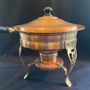 May include: A copper-colored chafing dish with a lid and a black handle. The dish sits on a brass-colored stand with decorative legs. The chafing dish is designed for keeping food warm.