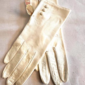 May include: A pair of off-white leather gloves featuring three decorative buttons on the wrist and a scalloped edge. The gloves include a tag marked with the number 7 and the brand name Grandoe, indicating washable leather construction.