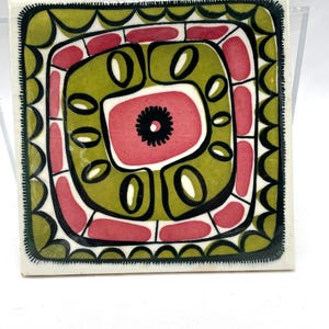 May include: Square ceramic tile with a colourful abstract design. The tile features a central pink shape with a black centre, surrounded by olive green and black shapes. The border is white with a scalloped edge.