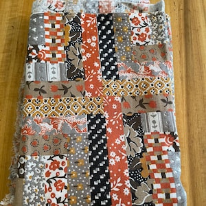 May include: Folded patchwork quilt featuring a variety of patterned fabrics in shades of orange, gray, brown, and white. The quilt has a rectangular shape with a fringed edge. The design includes floral, geometric, and abstract patterns.