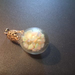 May include: A pendant featuring a small, clear glass globe filled with iridescent, multi-colored opal chips. The top of the glass globe is secured with an ornate, filigree-style metal cap in a warm, metallic bronze tone, featuring a small loop.