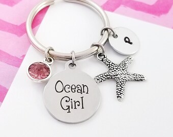 May include: A silver keychain with a pink gemstone, a starfish charm, and a round pendant that reads "Ocean Girl." A small, round charm with the letter "p" is also attached. The background is a pink and white chevron pattern.