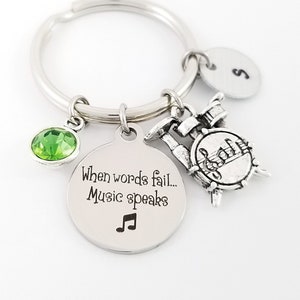 May include: Silver keychain with a green crystal charm, a drum charm, and a round charm with the text "When words fail... Music speaks" and a musical note symbol. The round charm also has a letter "S" on the back.