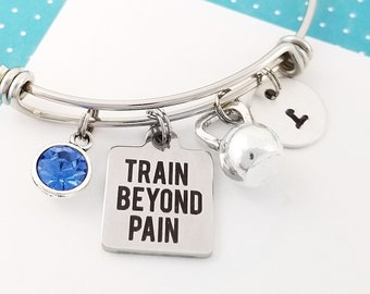 May include: Silver bangle bracelet with a blue crystal charm, a charm with the text "TRAIN BEYOND PAIN", and a charm shaped like a kettlebell. The bracelet also has a silver charm with the letter "L" on it.