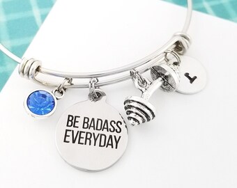 May include: Silver charm bracelet with a blue crystal charm, a dumbbell charm, and a round charm with the text "BE BADASS EVERYDAY".