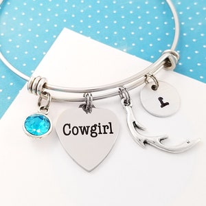 May include: Silver bangle bracelet with charms. The charms include a heart with the word "Cowgirl" on it, a small blue crystal, a small antler charm, and a round charm with the letter "L" on it.
