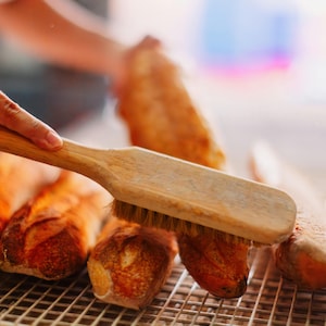 May include: A wooden brush with bristles is being used to brush a row of freshly baked baguettes on a wire rack.