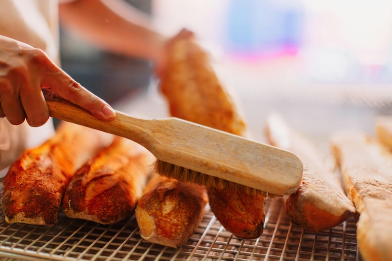 May include: A wooden brush with bristles is being used to brush a row of freshly baked baguettes on a wire rack.