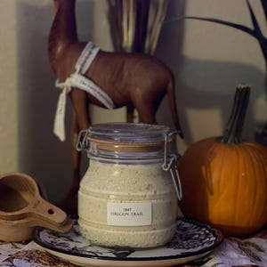 Peut inclure: Un bocal en verre avec une étiquette blanche sur laquelle est inscrit "1847 OREGON TRAIL" rempli d'une substance blanche. Le bocal est posé sur une assiette à motifs noirs et blancs sur une table avec une nappe à motifs marrons et blancs.