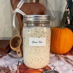 May include: A glass jar with a silver lid filled with a white, bubbly substance. The jar has a white label that reads "Gluten Free". The jar is sitting on a table with a brown wooden measuring cup and a pumpkin.