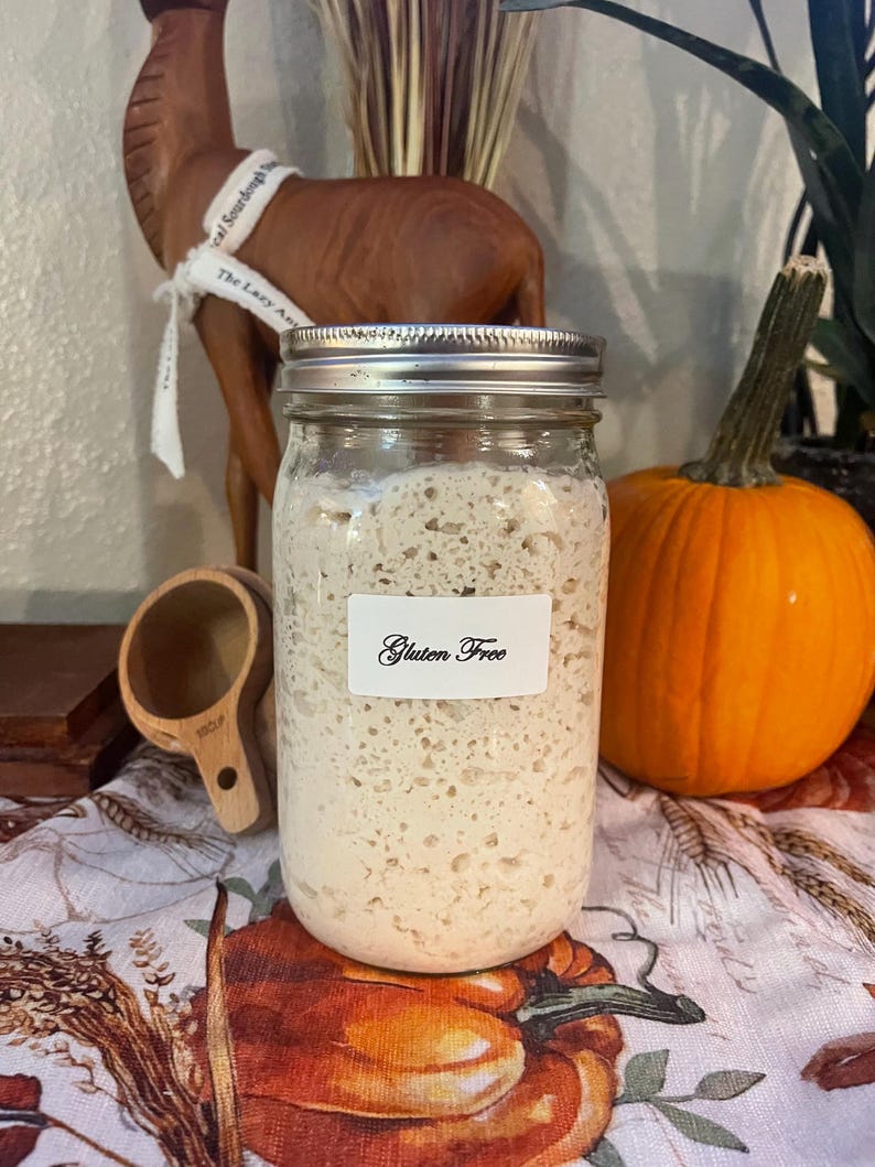 May include: A glass jar with a silver lid filled with a white, bubbly substance. The jar has a white label that reads "Gluten Free". The jar is sitting on a table with a brown wooden measuring cup and a pumpkin.