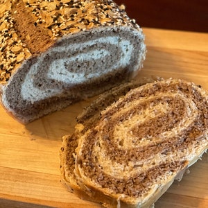 May include: A loaf of swirl bread with a brown crust and a swirl of white dough. The bread is topped with a variety of seeds, including sesame seeds, poppy seeds, and sunflower seeds. The bread is sliced and placed on a wooden cutting board.