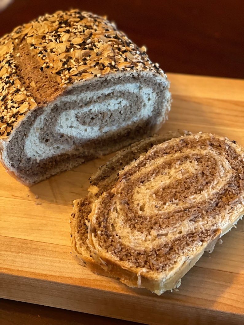 May include: A loaf of swirl bread with a brown crust and a swirl of white dough. The bread is topped with a variety of seeds, including sesame seeds, poppy seeds, and sunflower seeds. The bread is sliced and placed on a wooden cutting board.