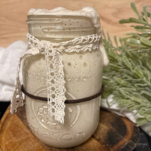 Op de afbeelding: Een glazen pot met een wit deksel en een bruin elastiekje om de bovenkant. De pot is gevuld met een witte, schuimende vloeistof. De pot is versierd met een wit kantlint.