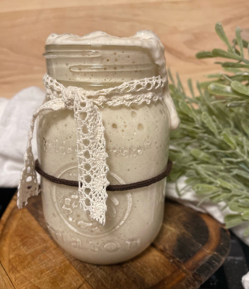 Op de afbeelding: Een glazen pot met een wit deksel en een bruin elastiekje om de bovenkant. De pot is gevuld met een witte, schuimende vloeistof. De pot is versierd met een wit kantlint.