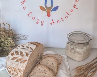 May include: A loaf of artisan bread with leaf-shaped scoring, sliced to reveal the interior texture. The bread rests on a wooden board with a white cloth. A jar with a clear lid and the text "The Lazy Antelope" on a white bag are also in the image.