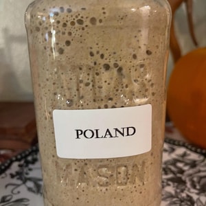 Puede incluir: Un frasco de vidrio con un líquido marrón en su interior. El frasco tiene una etiqueta blanca con la palabra "POLAND" escrita en ella.