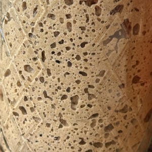 May include: Close-up of a glass jar filled with a light brown, bubbly mixture. The mixture has a porous texture with numerous air pockets and darker brown inclusions. The jar has a diamond pattern etched into the glass.